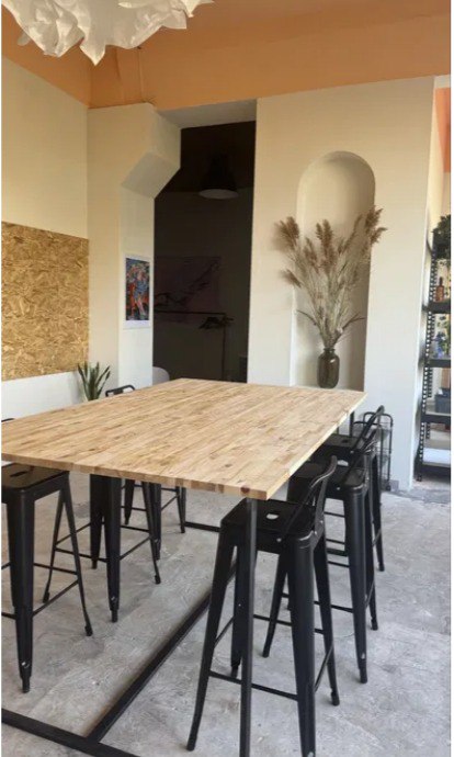 Bright and spacious Pigmentori workspace featuring a large raw wood table, black metal bar stools, wooden easels with paintings, dried pampas grass in a vase, and warm peach-beige walls